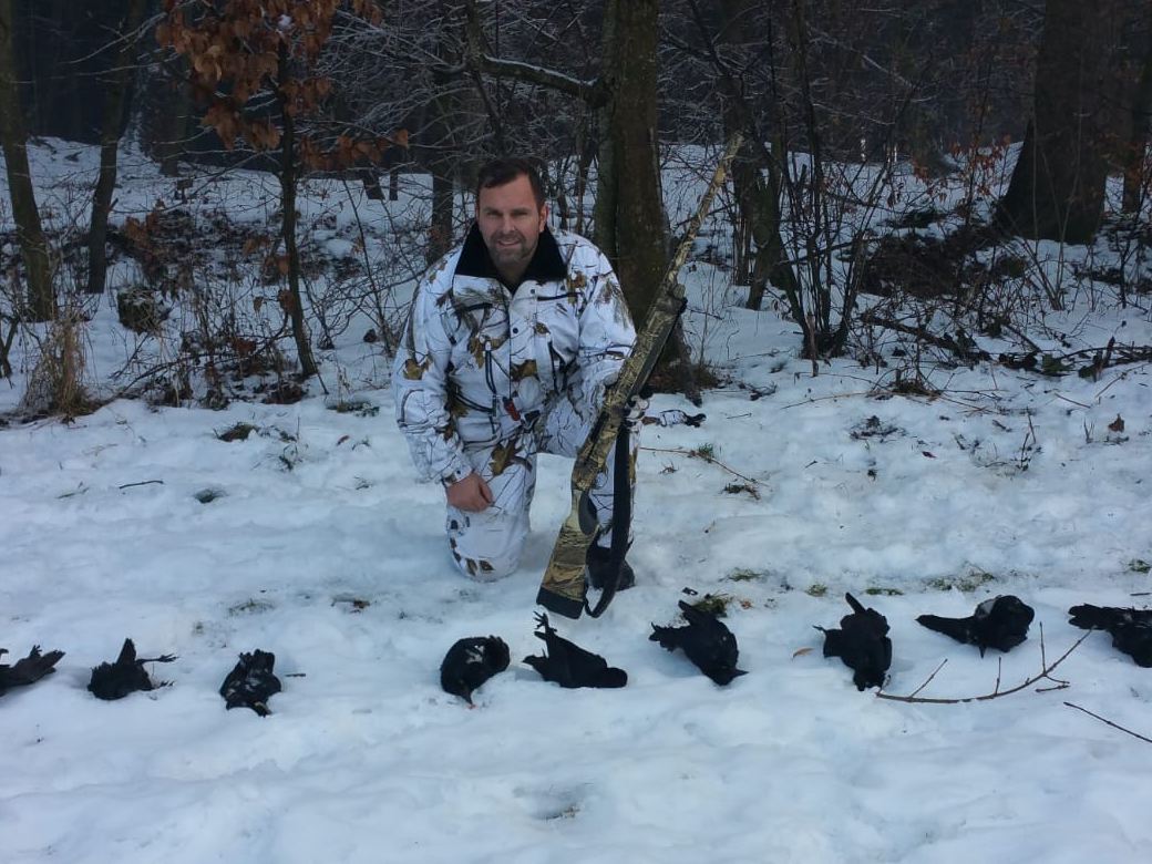 Christian Hanl in Schneetarnkleidung kniet mit Selbstladeflinte im verschneiten Wald, vor ihm eine Reihe erlegter Krähen